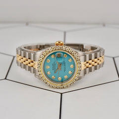 Front view of Rolex Ladies Datejust 26mm featuring ice-blue dial, diamond hour markers, and 18k yellow gold/steel bracelet on tiled surface.