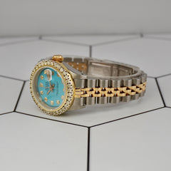 Angled side view of Rolex Ladies Datejust 26mm with 2ct diamond bezel, ice-blue dial, and two-tone Jubilee bracelet on geometric background.